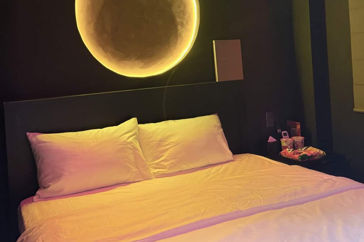 A bedroom with a large bed, a wall-mounted circular light, and a ceiling with a pink accent.