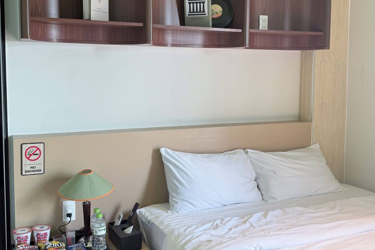 A hotel room with a bed, a nightstand, a shelf with books and a vase, and a 'No Smoking' sign.