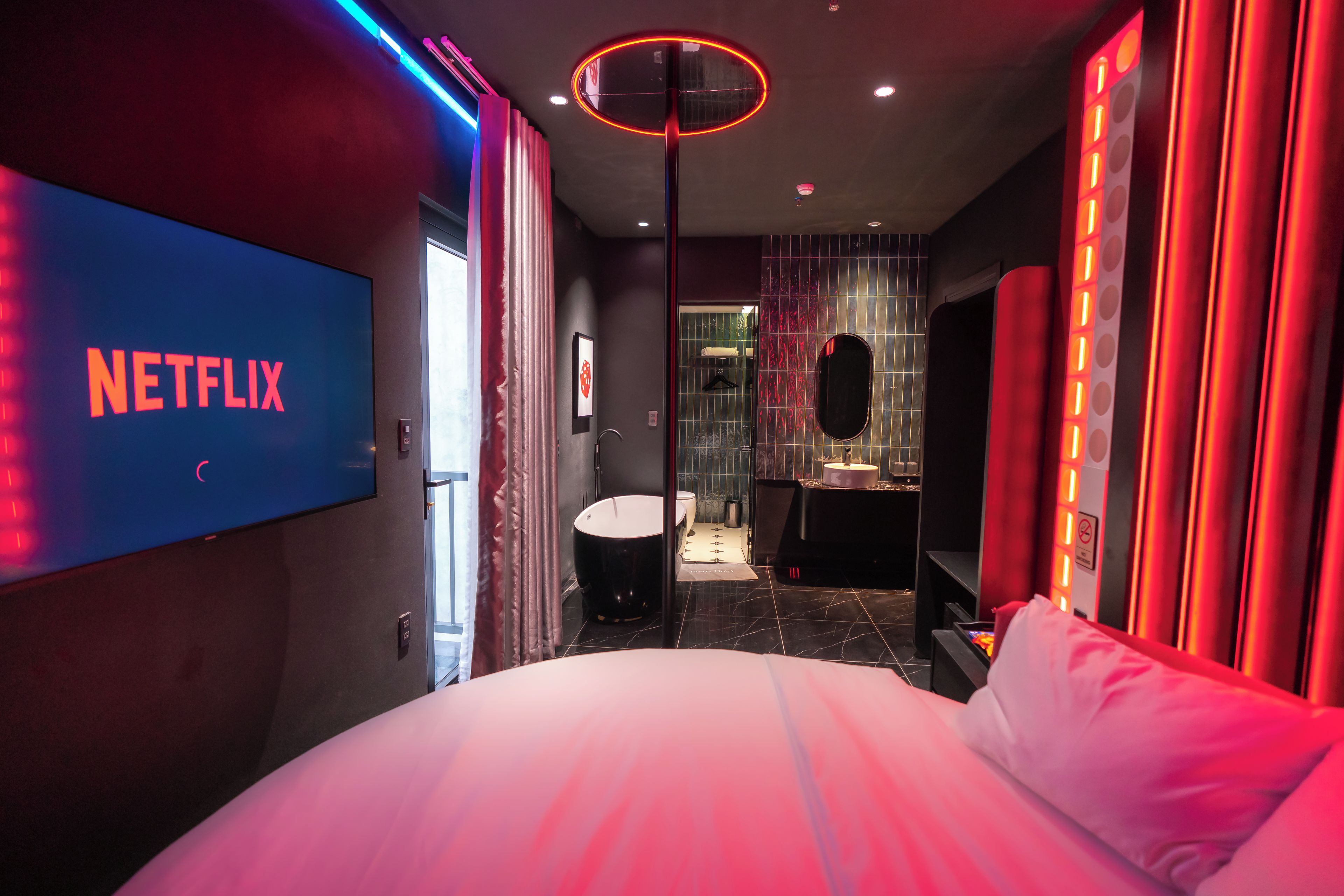 A modern hotel room with a large bed, a TV displaying the Netflix logo, and a bathroom with a mirror and sink.
