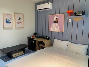 Habitación de hotel con cama, escritorio, refrigerador pequeño, aire acondicionado y obras de arte colgadas en la pared.