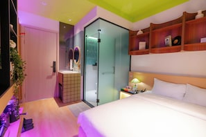 A hotel room with a bed, a bedside table with a lamp, a shelf with books and a bottle, a bathroom with a shower and toilet, and a door with a mirror.