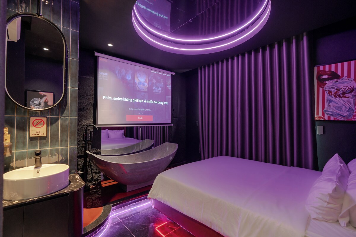 A modern hotel room with a large bed, a round bathtub, a vanity with a mirror, and a television screen displaying a menu.