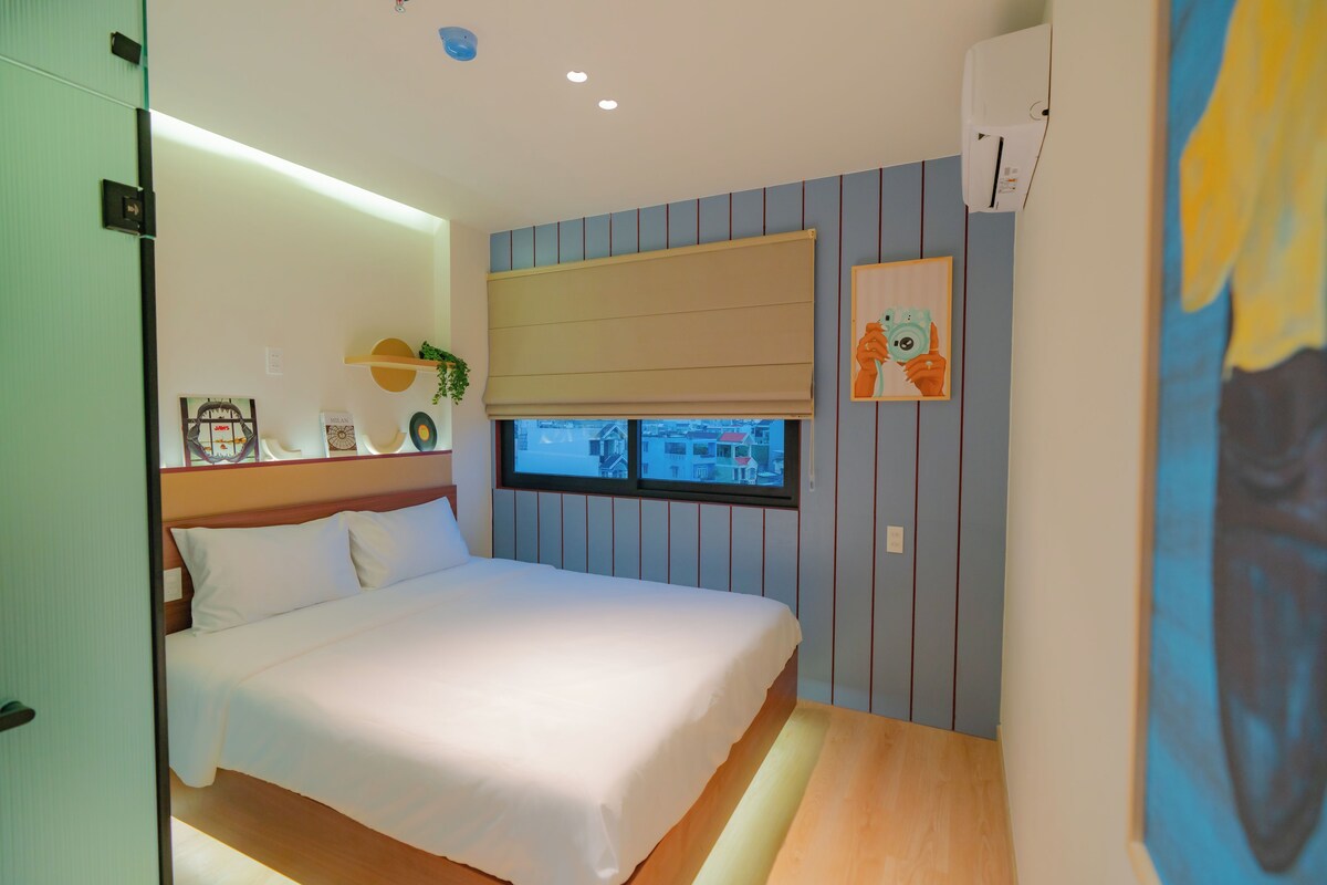 A bedroom with a bed, a window with a view, a shelf with decorative items, and a painting on the wall.
