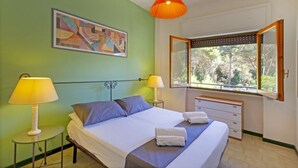 A bedroom with a bed, two bedside lamps, a window with a view, a painting on the wall, and a mirror above the window. - A cozy apartment with a balcony that sleeps up to four people, located within the Campoverde Residence in Vernole—set in a Mediterranean pine forest between San Cataldo and the Le Cesine State Nature Reserve. The property is just a short distance from  (Campo Verde)