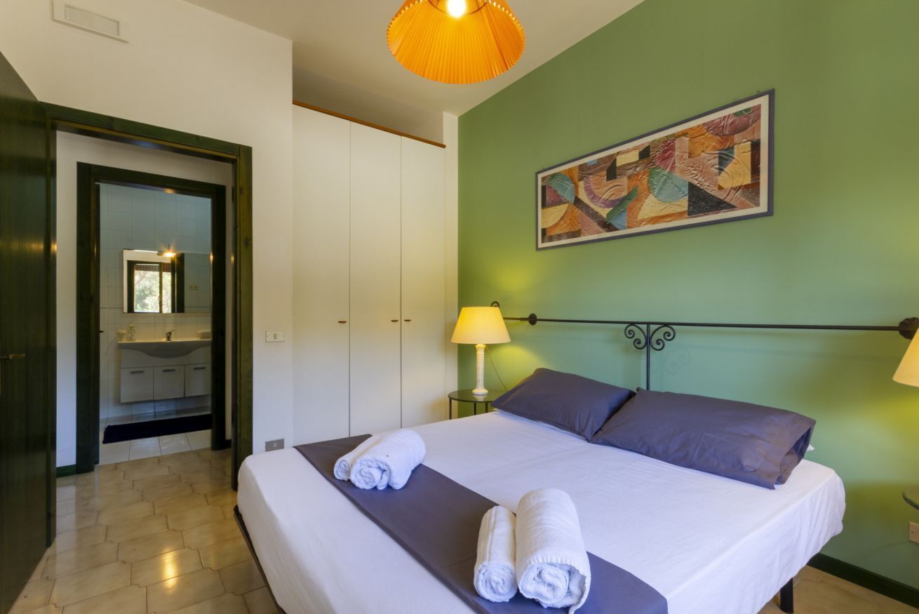 Habitación de hotel con cama, mesita de noche, lámpara, un cuadro y baño visible a través de una puerta abierta.
