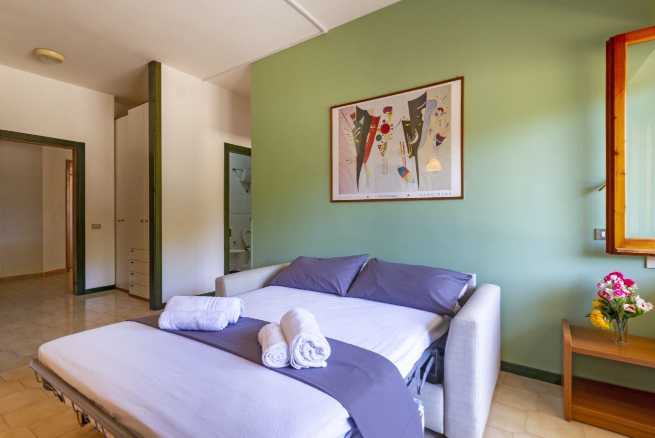 Een hotelkamer met een bed, kussens, een gekaderd kunstwerk aan de muur, een houten nachtkastje met een bloevas, en een deur naar een andere kamer.