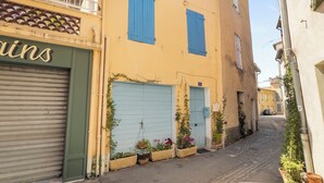 A narrow street with a yellow building, closed shutters, and potted plants.