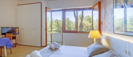 Habitación de hotel con cama, televisor, escritorio y ventana con vista a árboles.
