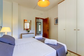 Hotellihuone, jossa on sänky, yöpöytä, lamppu, peili, vaatekaappi ja ovi, joka johtaa toiseen huoneeseen.