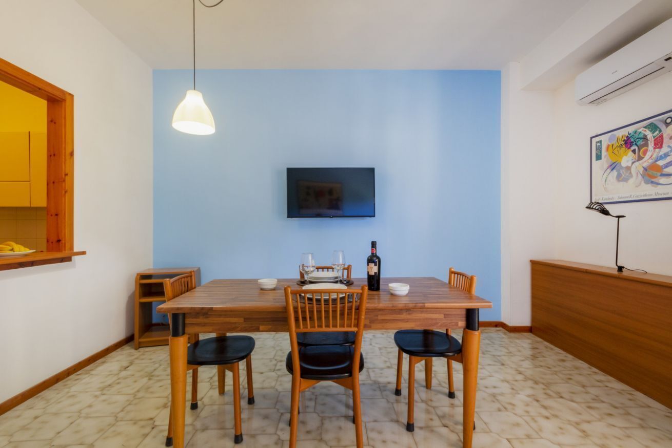 A dining area with a wooden table, four chairs, a television mounted on the wall, and a pendant light.