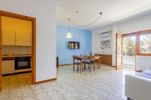 A compact kitchen with wooden cabinets, a dining area with a table and chairs, a hanging light fixture, and a view through a glass door. - A delightful apartment with a balcony that sleeps up to four people, located within the Campoverde Residence in Vernole—set in a Mediterranean pine forest between San Cataldo and the Le Cesine Nature Reserve. The property is just a short distance from  (Campo Verde)