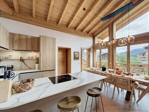 A modern kitchen with wooden cabinets, a marble countertop, and a dining area with a view. - The Fustic Apartment—Accommodates 8 People—Near the Slopes (Les Gets)