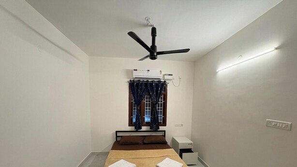 A small, clean room with a bed, a ceiling fan, an air conditioning unit, a window with curtains, and a bedside table. - The Value Stay Service Apartments (Chennai)