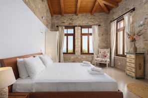 Un dormitorio con una cama grande, paredes de piedra, vigas de madera, una silla, un aparador y una ventana con cortinas.