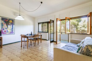 Ein Zimmer mit Esstisch, Stühlen, einer Couch, einem Gemälde und großen Fenstern mit Blick auf Bäume.