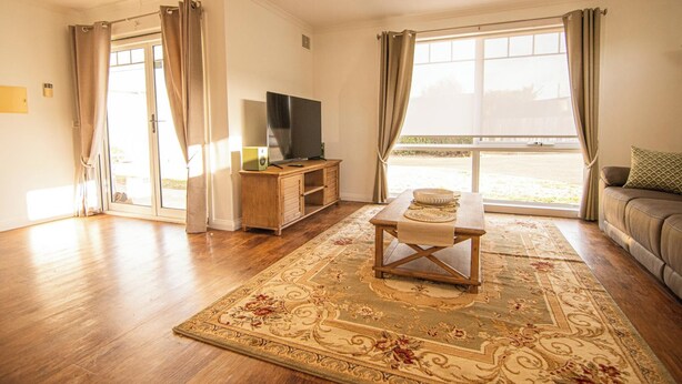 A living room with a wooden floor, a television on a wooden stand, a dining table with a table runner, a sofa with a cushion, and a large window with curtains.