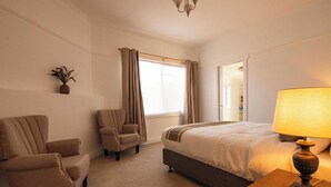 A hotel room with a bed, two armchairs, a lamp, a window with curtains, and a plant on the wall.