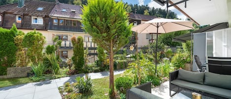 Un patio con mobiliario exterior, un jardín con diversas plantas y vistas a edificios residenciales al fondo.