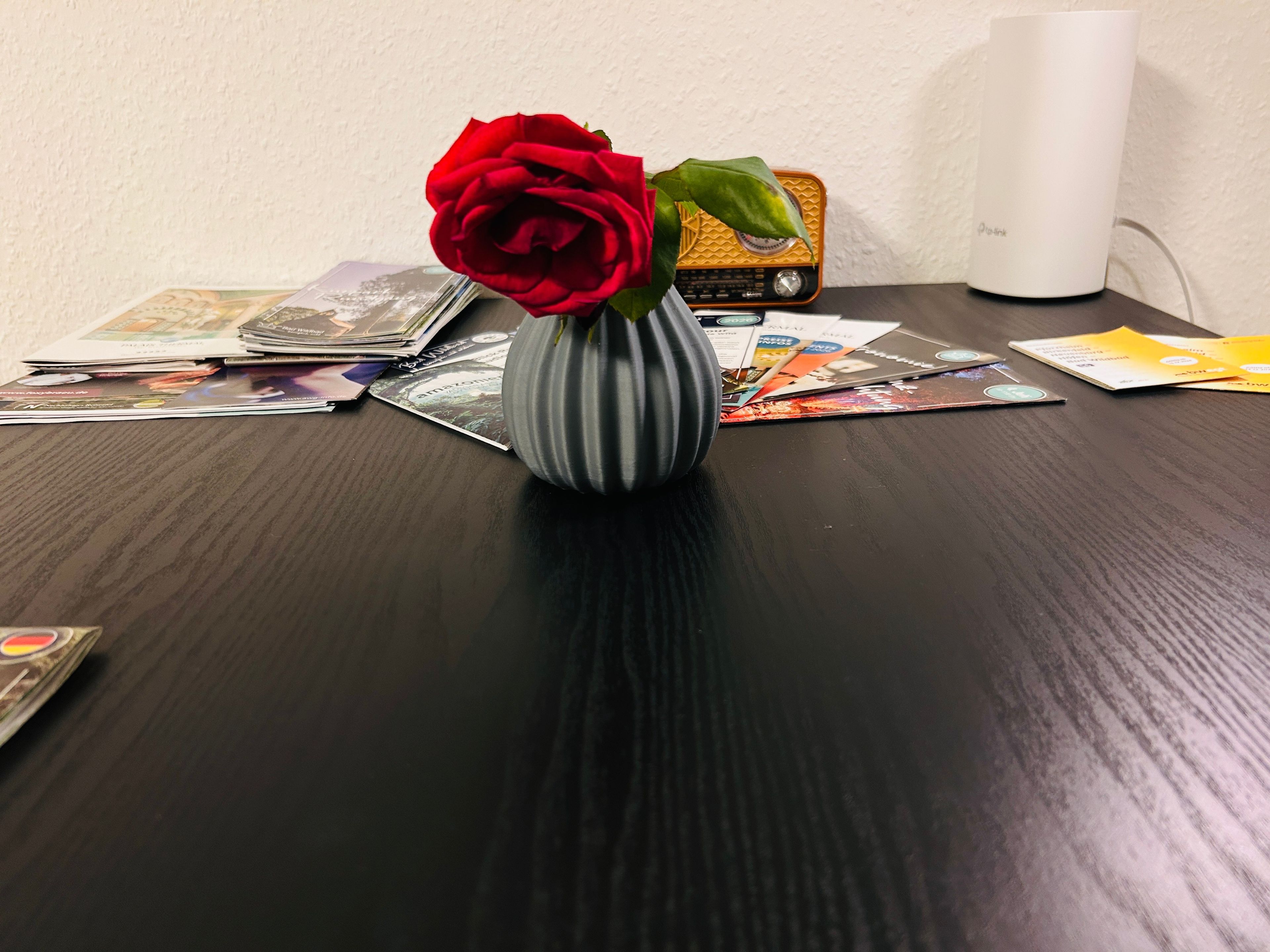 A table with a vase containing a single red rose, surrounded by various magazines and a vintage radio.