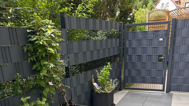 Ein Gartenbereich mit Fliesenboden, einem schwarzen Metallzaun und Kübelpflanzen.