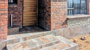 Un bâtiment en briques avec une porte en bois, une fenêtre protégée par une grille métallique et un petit escalier en pierre menant à l’entrée.