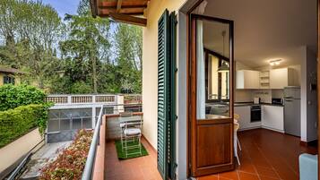 Un balcone con vista su un giardino e alberi, un piccolo tavolo con sedie e una cucina con pensili bianchi e un lavello.