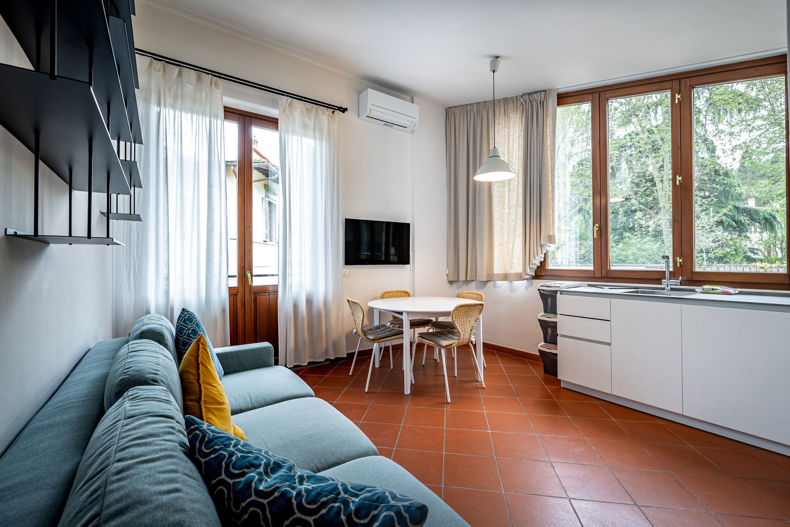 A modern living room with a sofa, a dining table with chairs, a kitchen area with a sink and cabinets, and a large window with curtains.