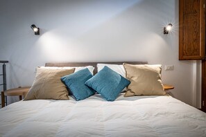 En snyggt bäddad säng med ett vitt täcke och två kuddar, en blå och en beige, i ett rum med en sänggavel i trä och en väggmonterad lampa.