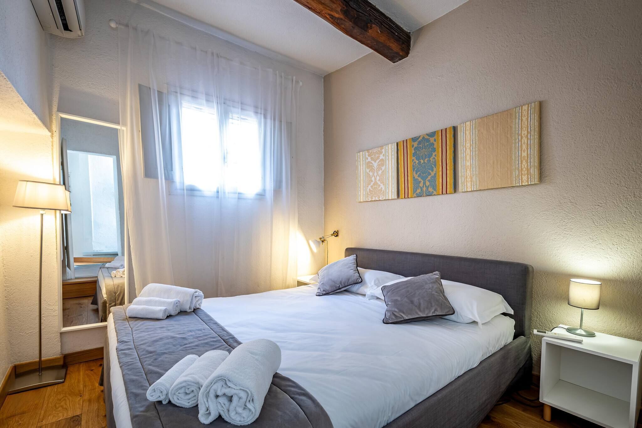 Ein ordentlich bezogenes Bett mit weißen Leinen und grauer Bettwäsche, ein Nachttisch mit Lampe sowie ein an der Wand hängendes Kunstwerk.