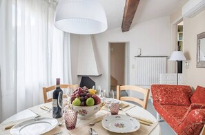 Ein Esstisch ist gedeckt mit Tellern, Besteck, einer Obstschale und einer Weinflasche. Im Raum befindet sich eine rot gemusterte Couch und eine weiße Lampe.