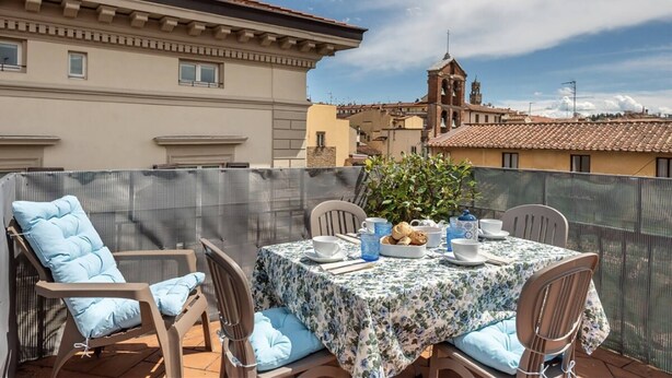 Terraço no telhado com uma mesa posta para dois, cercado por cadeiras, com vista para uma paisagem urbana com edifícios e uma torre de igreja.