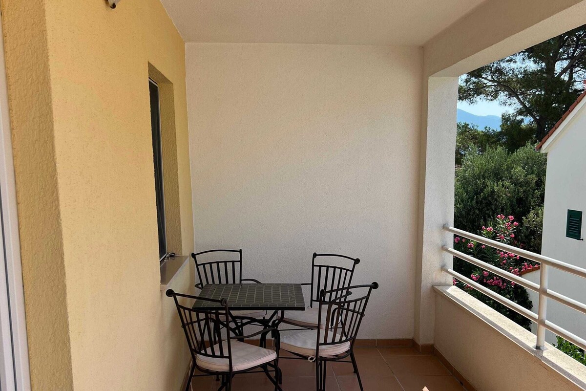 Ein kleiner Balkon mit Tisch und Stühlen, ein Fenster und Blick ins Grüne.