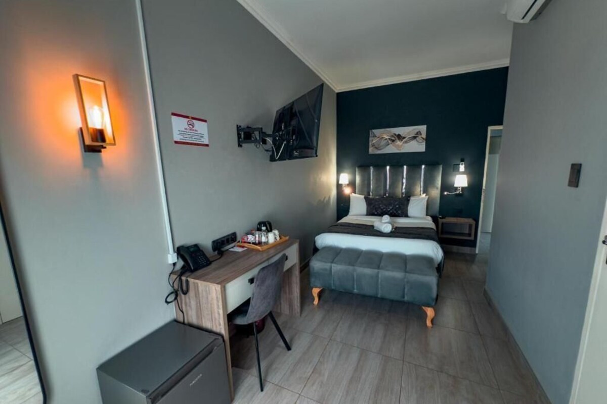 A compact hotel room with a bed, bedside table, desk, chair, and a small refrigerator.