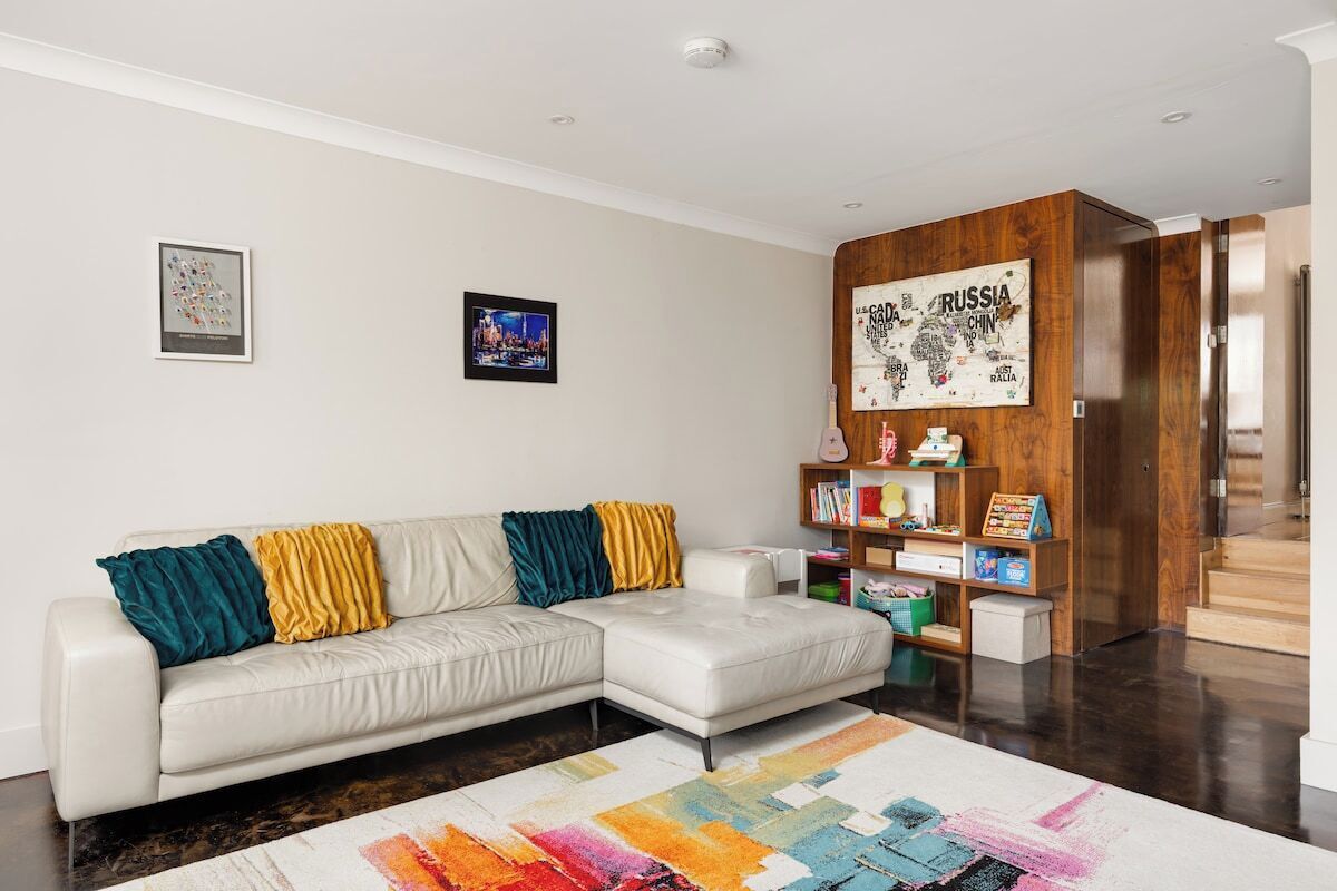 A modern living room with a white sectional sofa, colorful cushions, a wooden bookshelf, and a large rug.