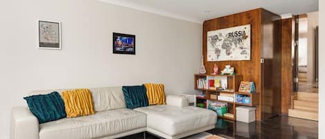 A modern living room with a white sectional sofa, colorful cushions, a wooden bookshelf, and a large rug.