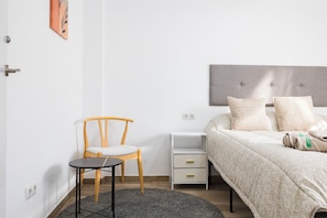 Een slaapkamer met een bed, een stoel, een klein tafeltje, een nachtkastje en een kunstwerk aan de muur.