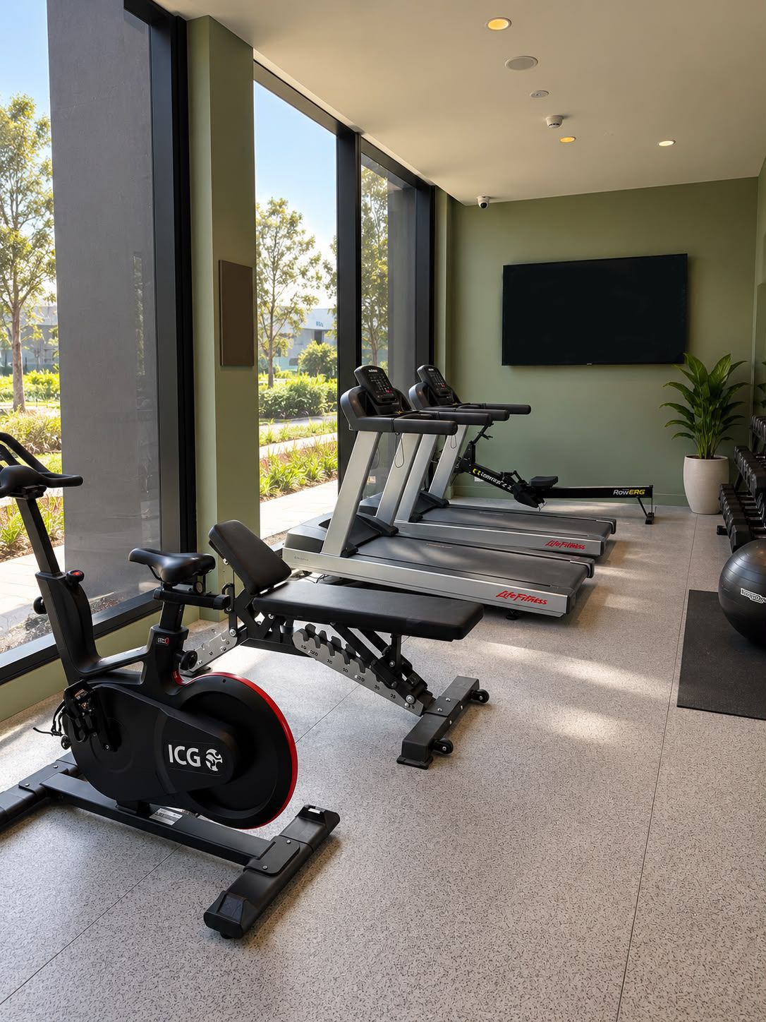 A gym with cardio machines, weight benches, and a large window offering a view of the outdoors.