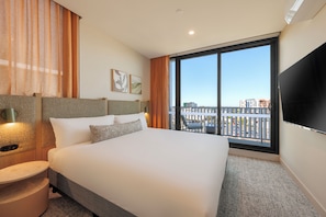 Habitación de hotel con una cama grande, televisor de pantalla plana, vista panorámica de la ciudad y balcón con mobiliario exterior.