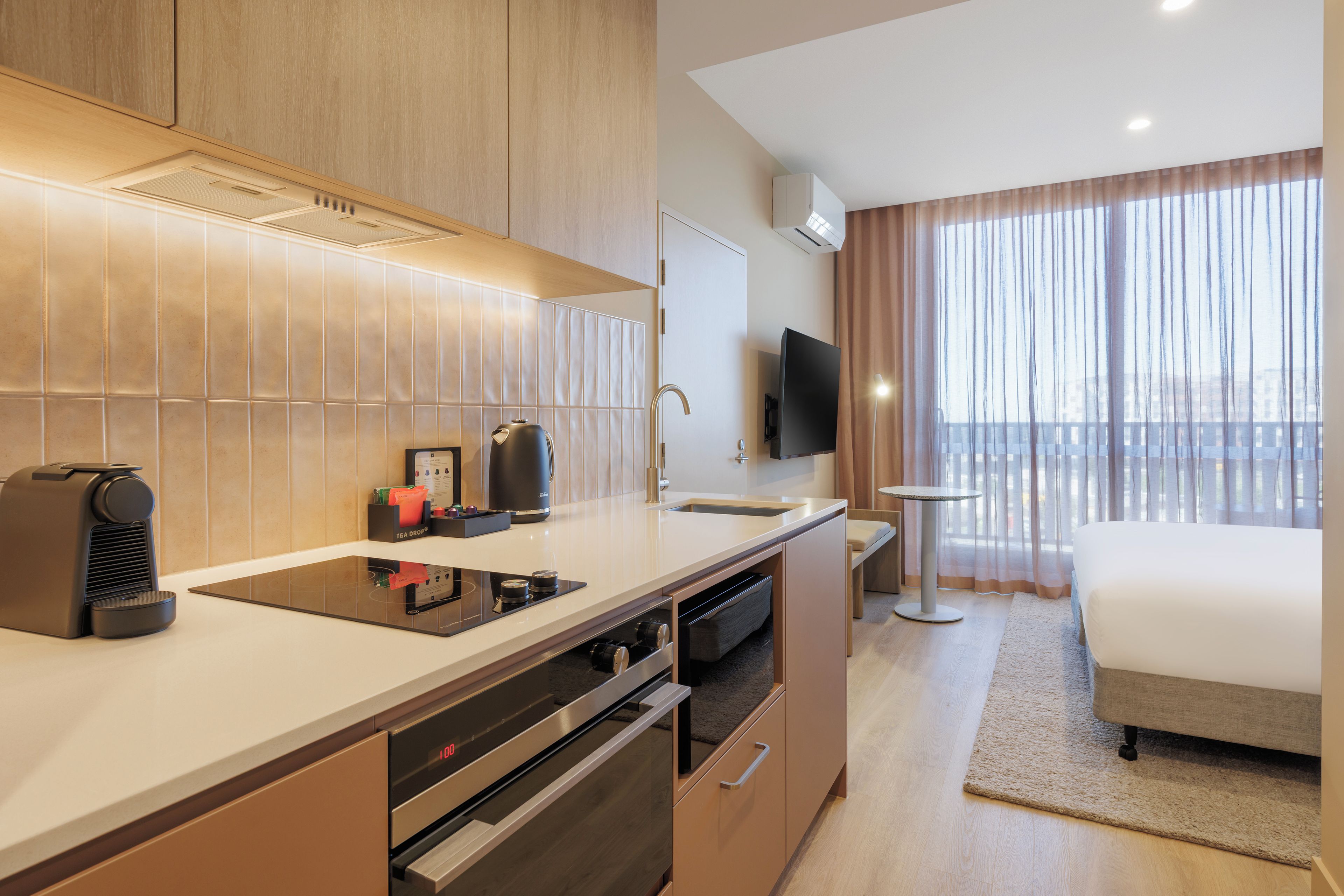 A modern hotel room with a kitchenette, a bed, a TV, and a view of the city.