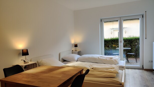 Une chambre avec un lit en bois, des tables de chevet, un bureau et une chaise. Une porte-fenêtre coulissante donne accès à un espace extérieur aménagé avec une table et des chaises.