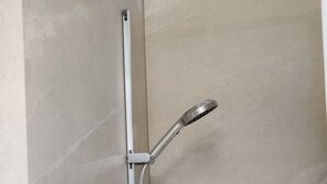 Une douche avec un pommeau de douche mobile fixé au mur, un panneau de commande situé en dessous et un mur recouvert de carreaux.