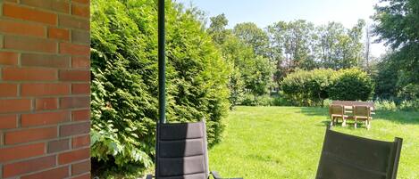 A patio area with a brick wall, a table, and chairs, surrounded by a grassy lawn and dense foliage.