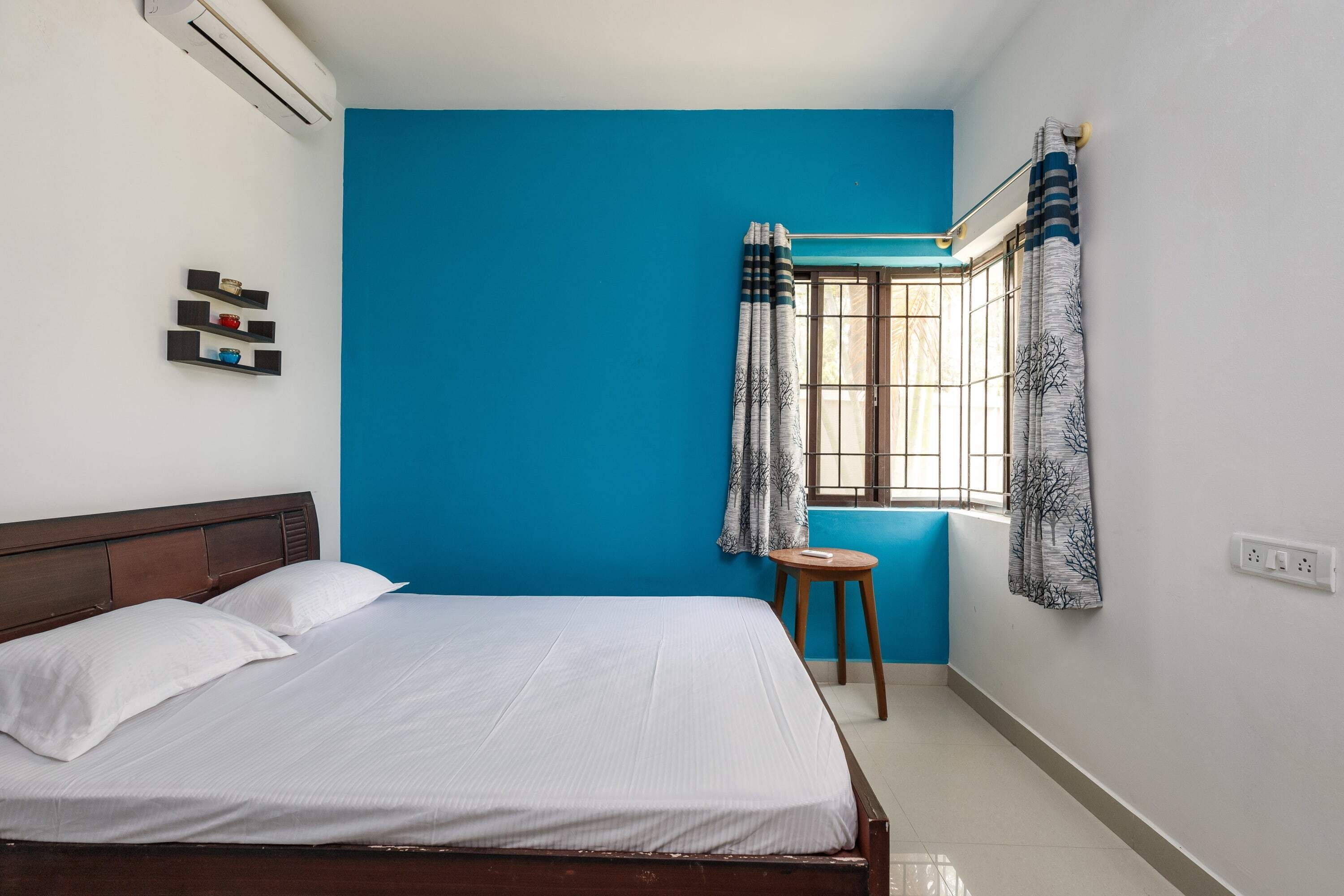 A room with a bed, a stool, a window with curtains, and an air conditioning unit.