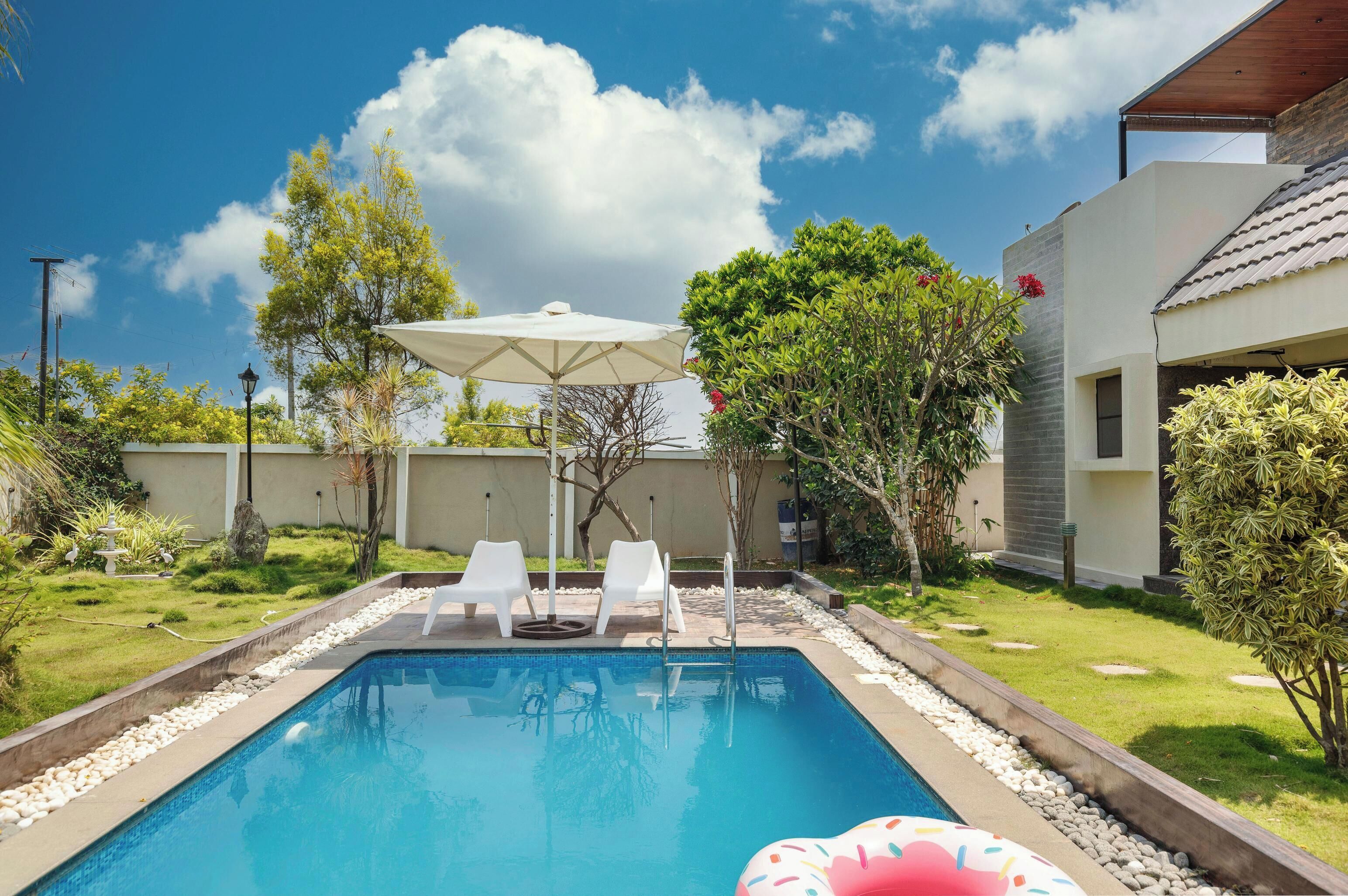 Une piscine entourée de verdure, avec des chaises longues blanches et un parasol, sous un ciel dégagé.