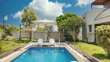 Une piscine entourée de verdure, avec des chaises longues blanches et un parasol, sous un ciel dégagé.