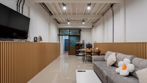 A modern living room with a flat-screen TV, a grey sofa with a large flower pillow, a white ottoman, and a wooden partition. - Cool Cozy 3-bedroom townhome in Chinatown Krung Thep Maha Nakhon (Krung Thep Maha Nakhon)