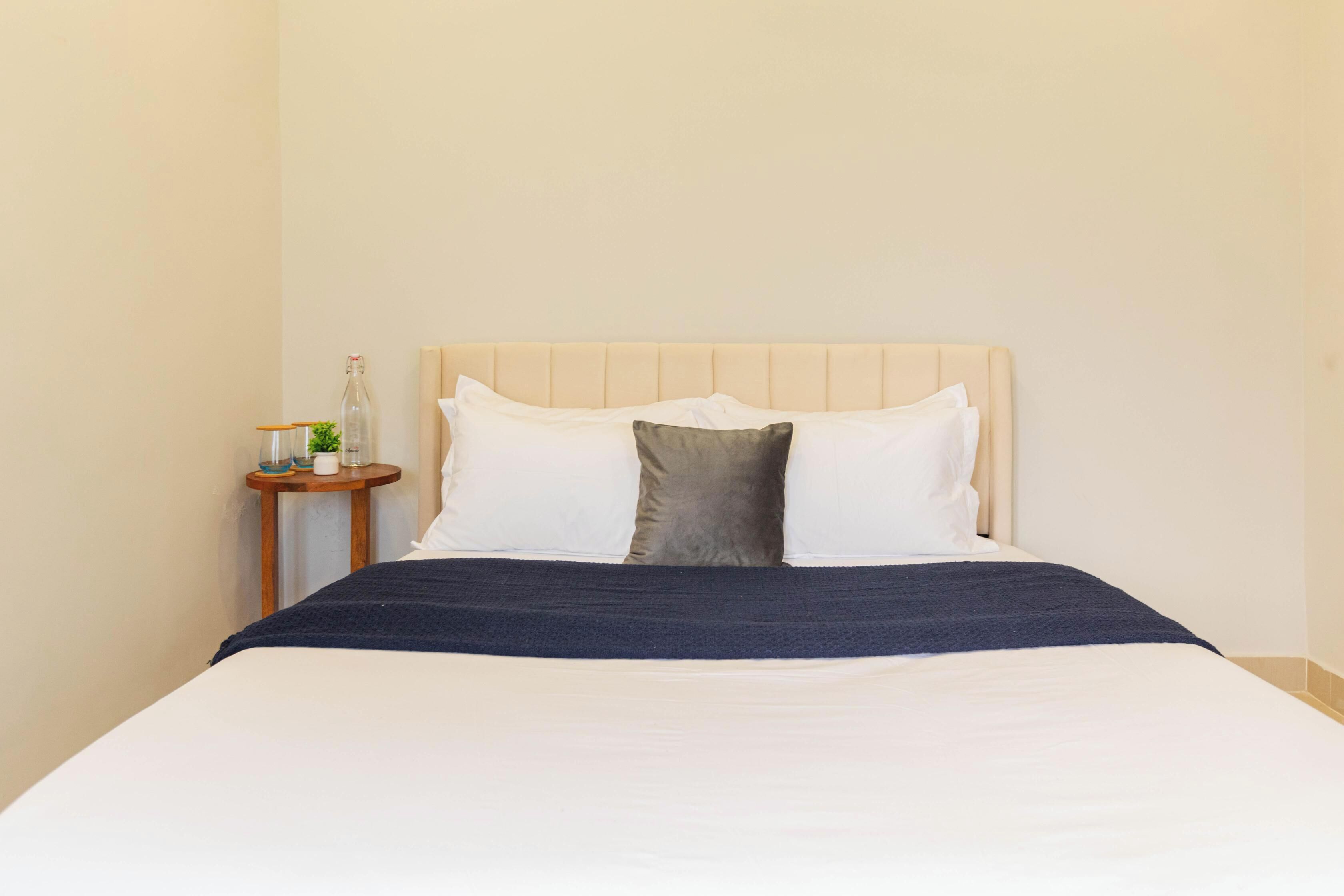 Una cama bien hecha con cubrecama blanco y azul oscuro, una mesita de noche de madera con una planta pequeña y un cabecero color beige.