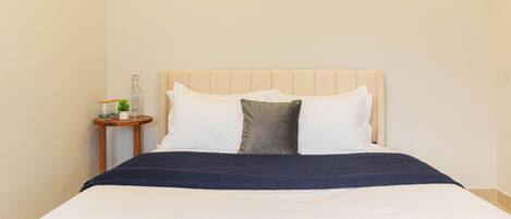 Una cama bien hecha con cubrecama blanco y azul oscuro, una mesita de noche de madera con una planta pequeña y un cabecero color beige.