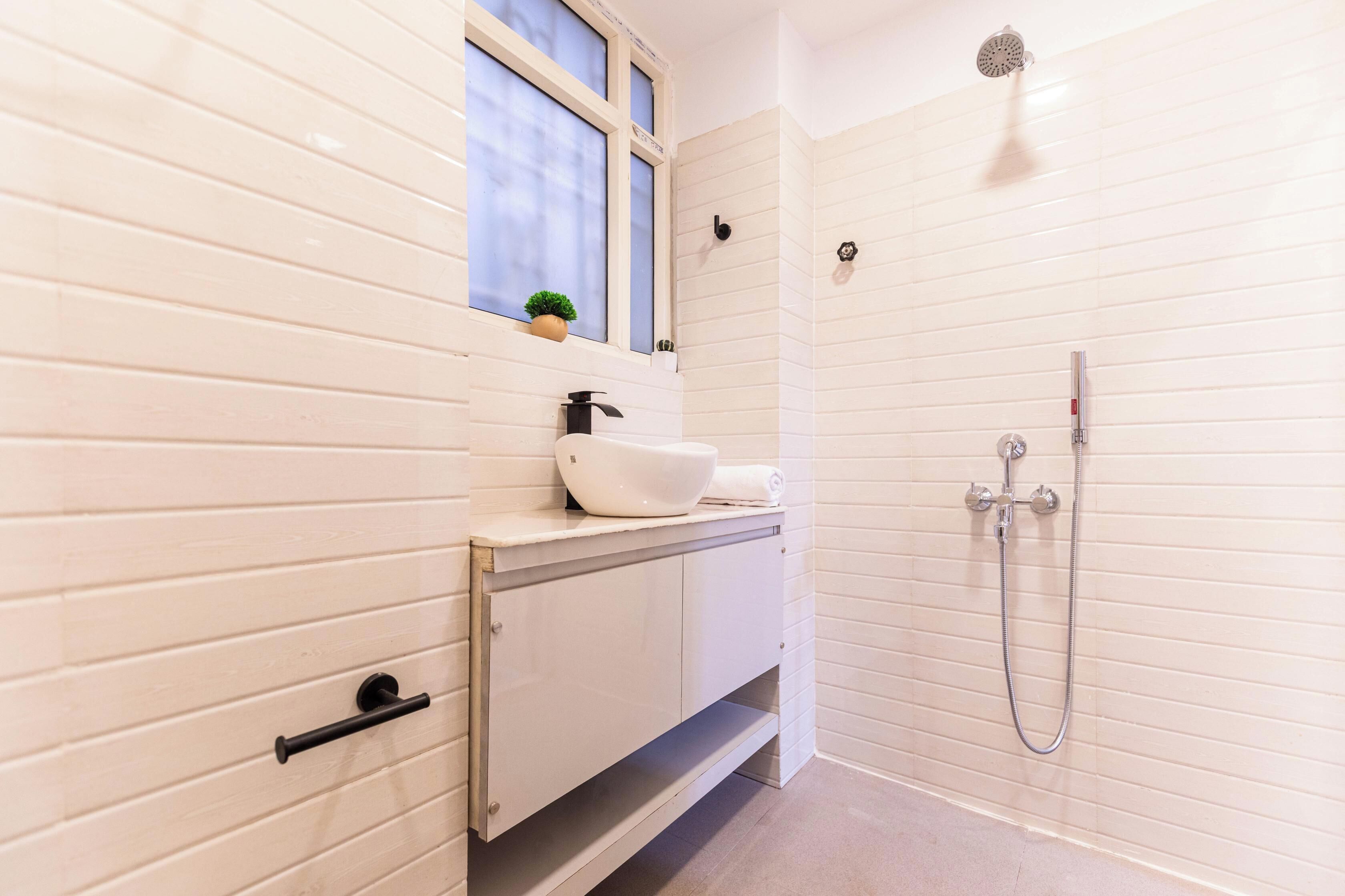 A bathroom with a white tiled wall, a sink with a faucet, a small window with a plant, and a shower area with a handheld showerhead.