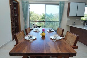 A dining table with chairs, a yellow teapot, and a view of greenery through a large window. - Aurora Mansion by Red Olive 3bhk Nr ECR Beach (Chennai)